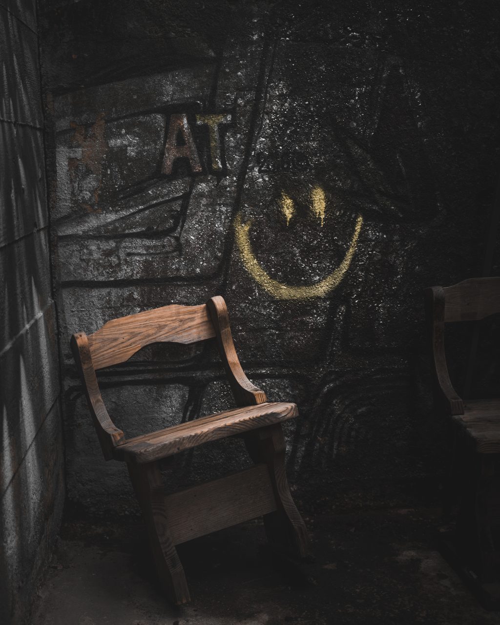 A wooden chair against a wet concrete wall with a yellow smiling face painted above it.