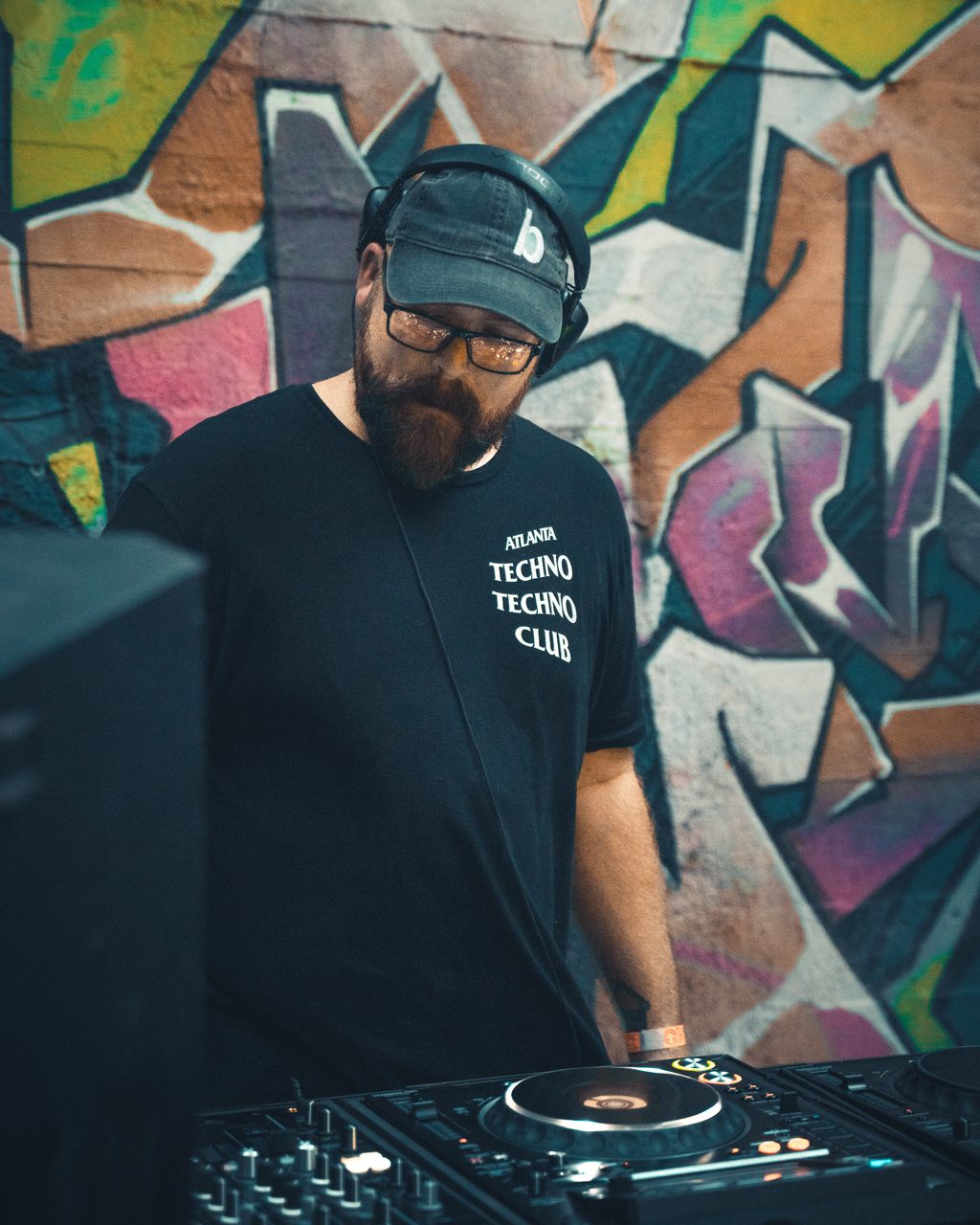 The performer standing over the mixer in a cap and headphones, graffiti filling the frame behind him.