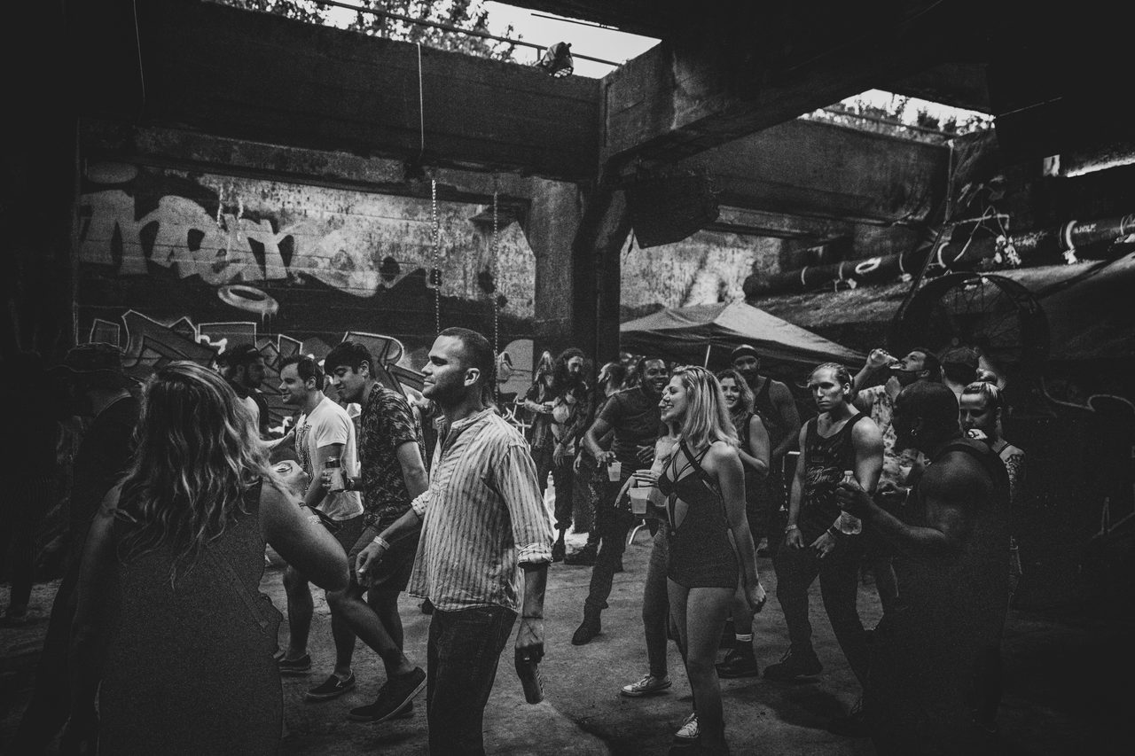 Black and white. A crowd standing and talking under a concrete overpass.