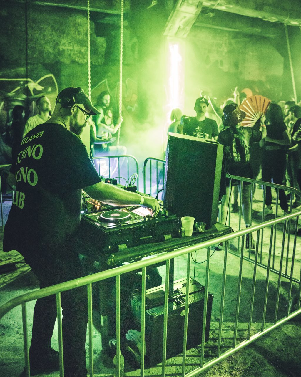The performer at the decks behind a metal barrier, the room flooded green from a light above the crowd.