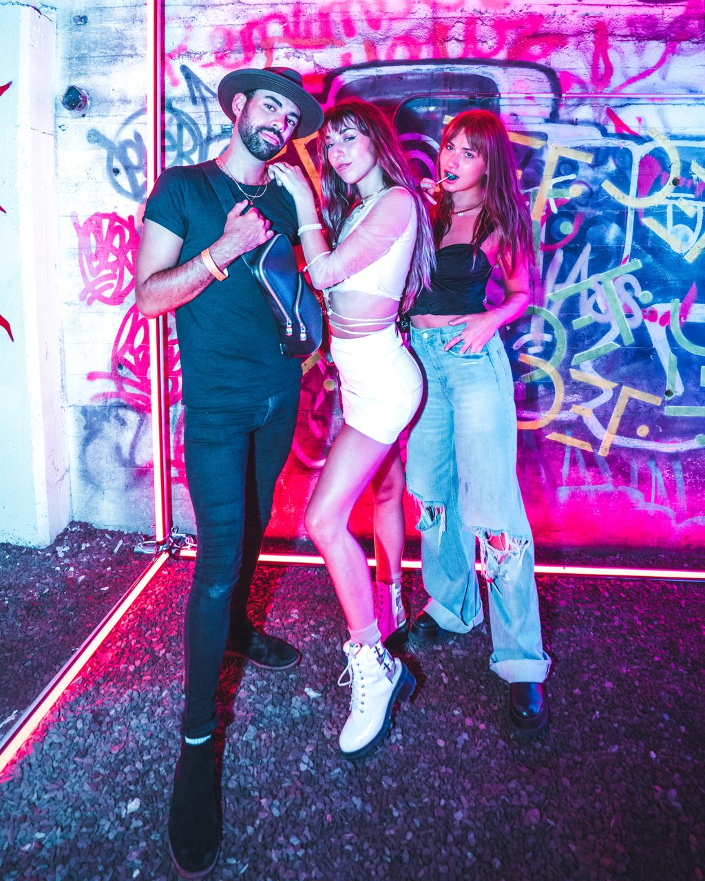 Three people against a graffiti wall lit magenta, a neon tube running along the floor behind them.