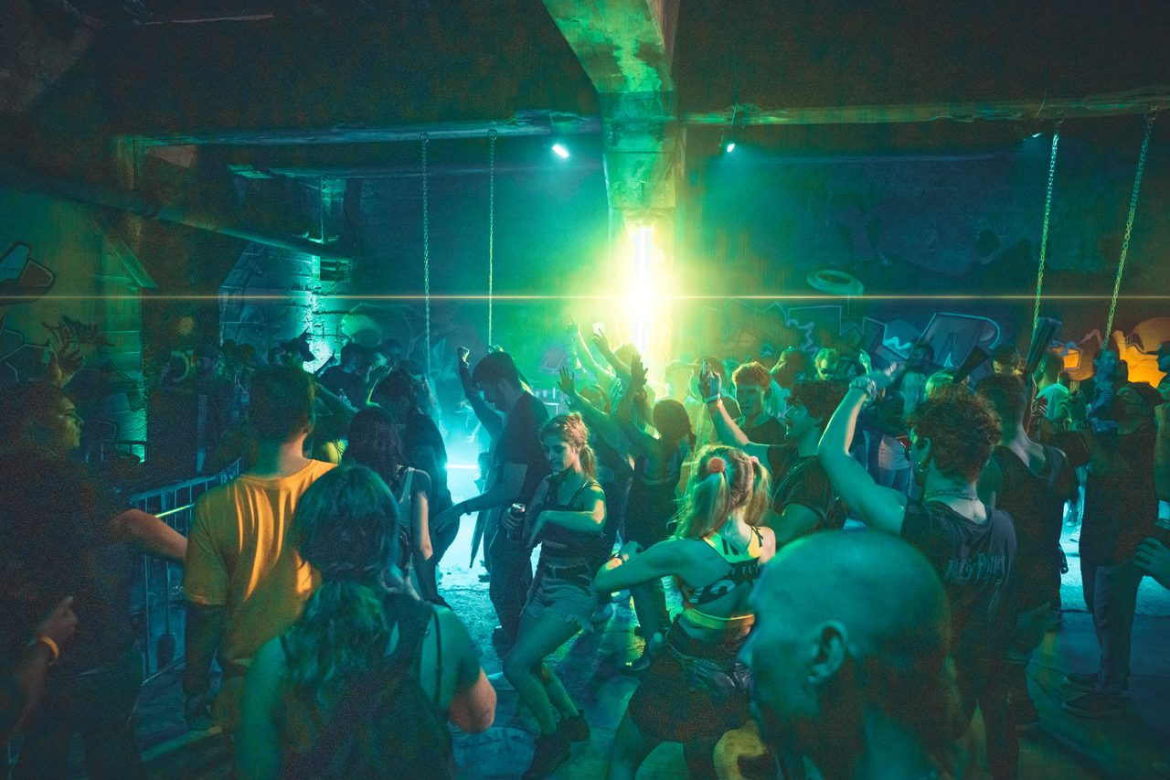 A crowd dancing under a green beam that fills the ceiling of a concrete room.