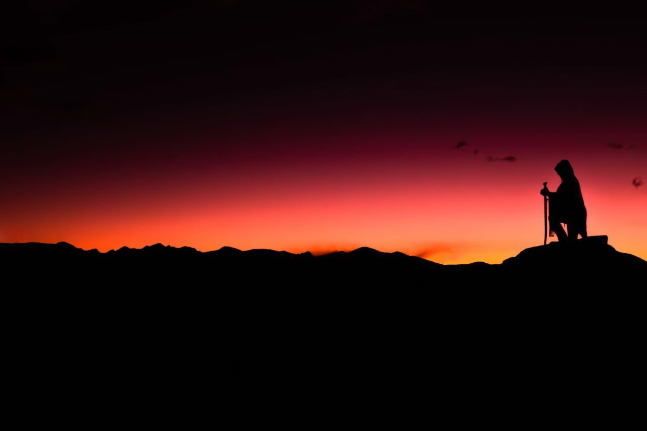 A kneeling figure with a sword on a ridge against a crimson sky.